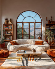 Modern living room with a large sofa, striped orange Moroccan rug, coffee table, and bookshelves with a scenic window view.