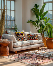 Living room with a pink Moroccan rug, white sofa, patterned cushions, and large windows with city view.